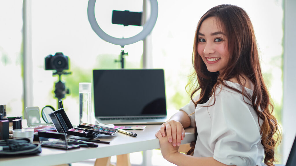 Young and cute Asian female vlogger, influencer, or online seller sitting with cosmetics products and DSLR camera and smartphone and laptop notebook computer ready to broadcast video live stream